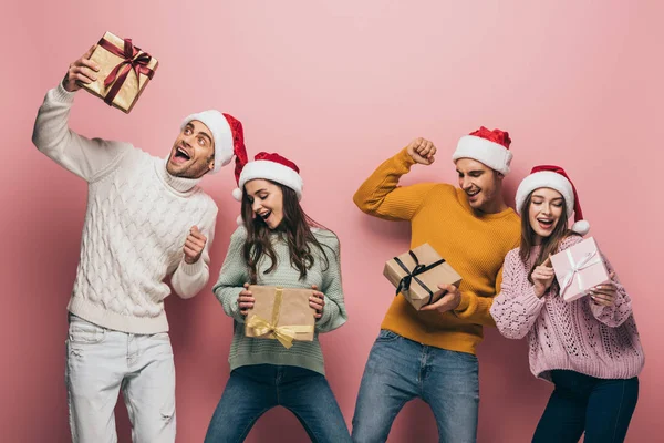Amigos animados em chapéus santa segurando presentes de Natal, isolado em rosa — Fotografia de Stock