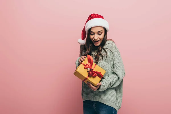 Menina surpreso em suéter e chapéu de santa segurando presente de Natal, isolado em rosa — Fotografia de Stock