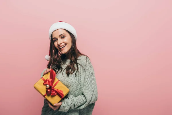 Smiling girl in sweater and santa hat holding christmas gift, isolated on pink — Stock Photo