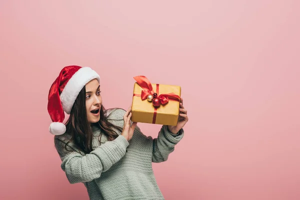 Donna sorpresa in maglione e cappello di Babbo Natale in possesso di regalo di Natale, isolato su rosa — Foto stock