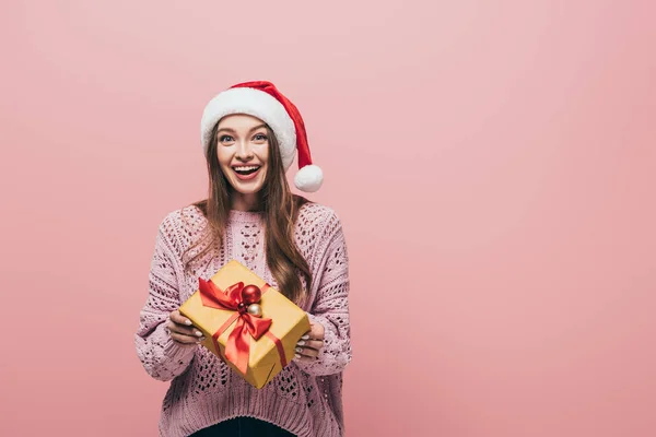 Lächelnde Frau in Pullover und Weihnachtsmütze mit Weihnachtsgeschenk, isoliert auf rosa — Stockfoto