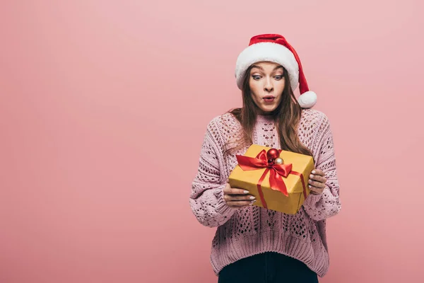 Überraschte Frau in Pullover und Weihnachtsmütze mit Weihnachtsgeschenk, isoliert auf rosa — Stockfoto