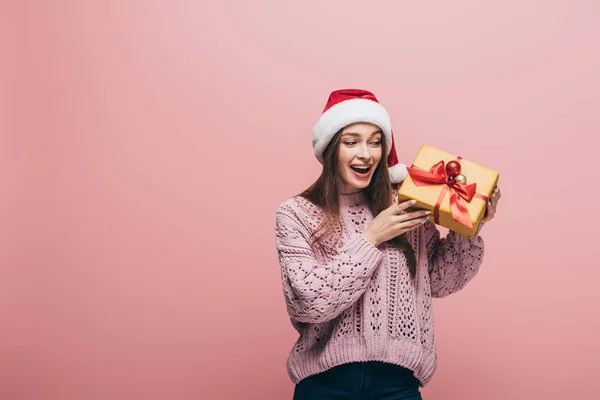 Excited woman in sweater and santa hat holding christmas gift, isolated on pink — Stock Photo