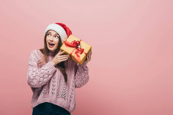 Emotional woman in sweater and santa hat holding christmas gift, isolated on pink — Stock Photo