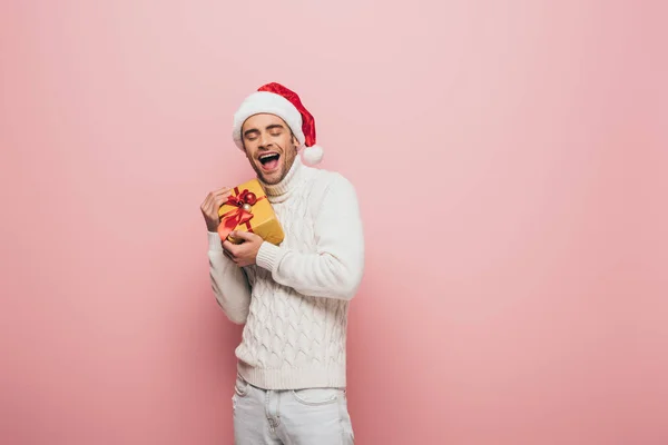 Excited man in santa hat holding christmas gift, isolated on pink — Stock Photo