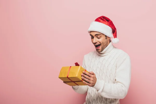 Aufgeregter Mann in Pullover und Weihnachtsmütze mit Weihnachtsgeschenkbox, isoliert auf rosa — Stockfoto