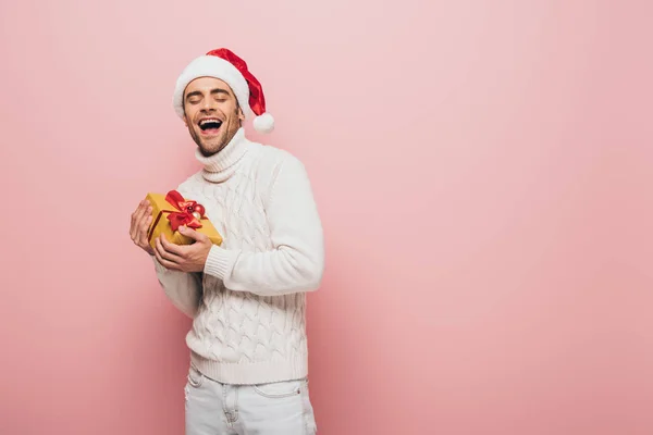 Excité bel homme en pull et chapeau santa tenant boîte cadeau de Noël, isolé sur rose — Photo de stock