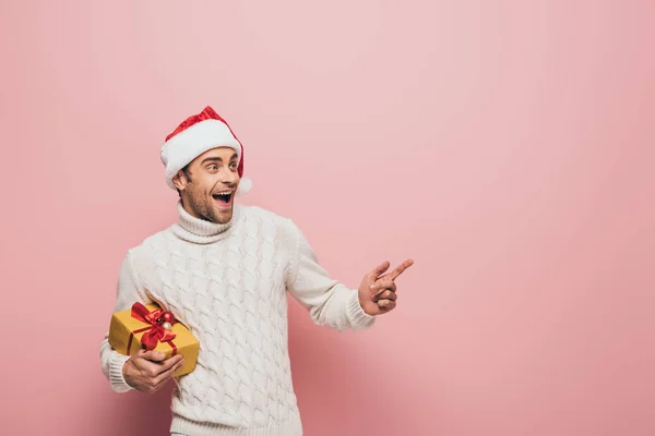 Handsome excited man in santa hat pointing and holding christmas present, isolated on pink — Stock Photo