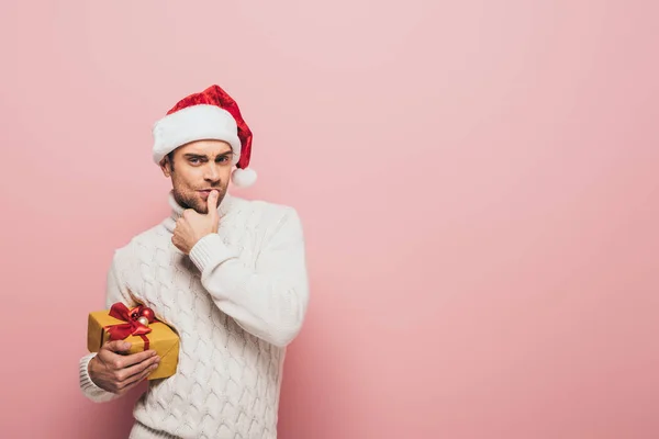 Thoughtful man in sweater and santa hat holding christmas present, isolated on pink — Stock Photo