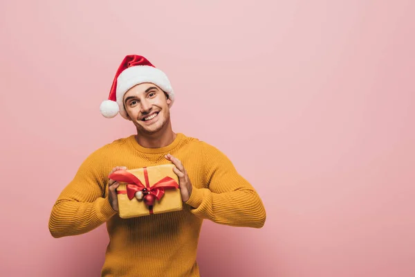 Handsome smiling man in sweater and santa hat holding christmas gift box, isolated on pink — Stock Photo