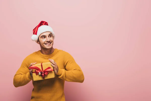 Smiling man in sweater and santa hat holding christmas gift box, isolated on pink — Stock Photo