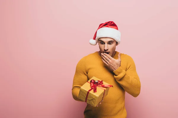 Schockierter Mann in Pullover und Weihnachtsmütze blickt auf Weihnachtsgeschenk, isoliert auf rosa — Stockfoto