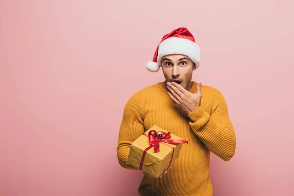 Шокований чоловік у капелюсі Санти жестикулює і тримає подарунок Різдва, ізольований на рожевому — стокове фото