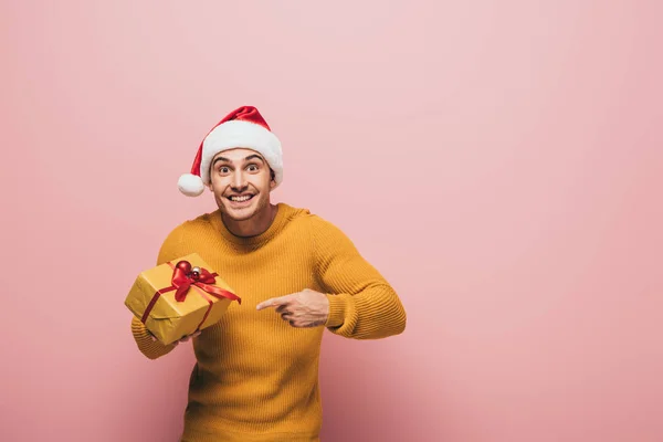 Fröhlicher Mann in Pullover und Weihnachtsmütze, der auf ein Weihnachtsgeschenk zeigt, isoliert auf rosa — Stockfoto