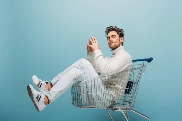 Handsome pensive man sitting in shopping cart, isolated on blue — Stock Photo