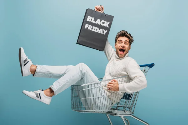 Excited man sitting in shopping cart while holding shopping bag with black friday sign, isolated on blue — Stock Photo