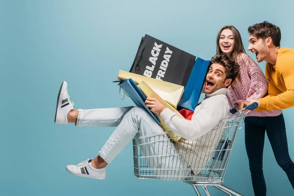 Amigos animados se divertindo com carrinho de compras e sacos de compras na sexta-feira preta, isolado em azul — Fotografia de Stock