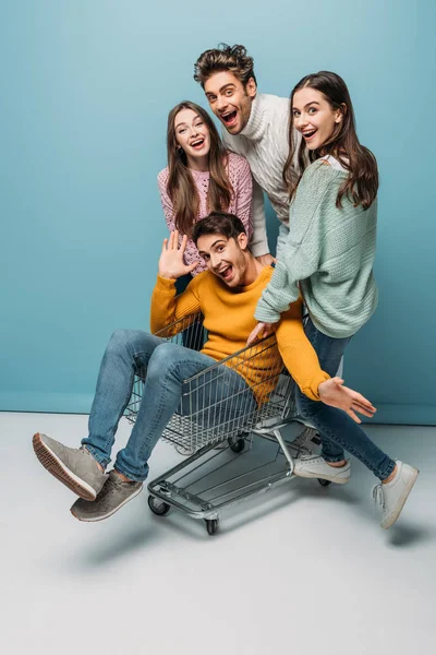 Happy friends having fun and riding in shopping cart on blue — Stock Photo