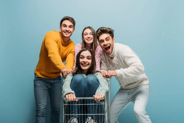 Amigos emocionados divirtiéndose con el carrito de compras, aislado en azul - foto de stock