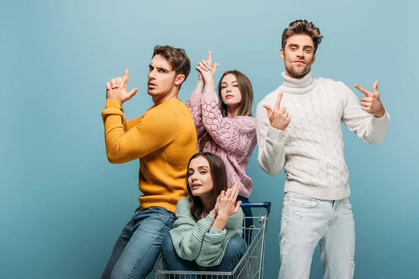 Happy friends making hand guns and having fun with shopping cart, isolated on blue — Stock Photo