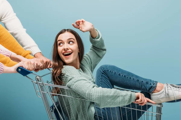 Amigos se divertindo e montando menina emocional no carrinho de compras, isolado em azul — Fotografia de Stock