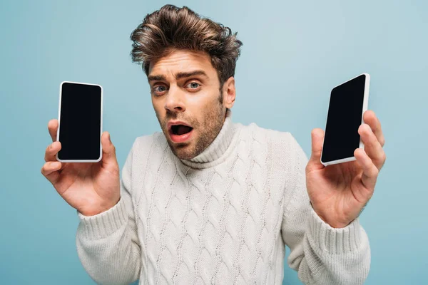 Hombre estresado mostrando dos teléfonos inteligentes con pantallas en blanco, aislado en azul - foto de stock