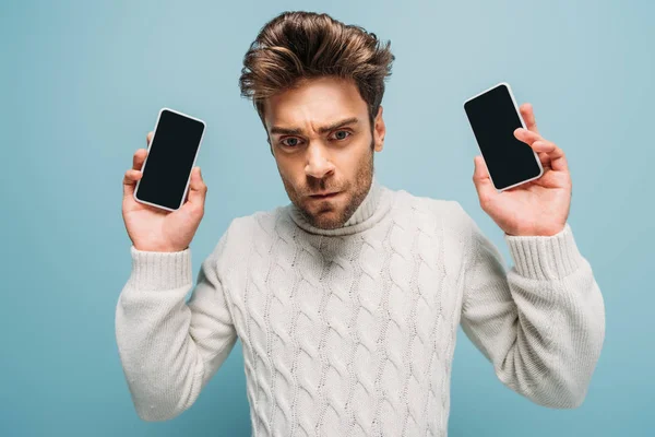 Hombre molesto mostrando dos teléfonos inteligentes con pantallas en blanco, aislado en azul - foto de stock