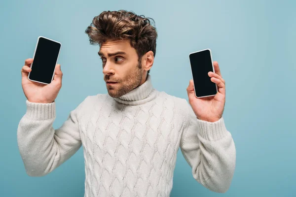 Emotionaler Mann blickt auf zwei Smartphones mit leeren Bildschirmen, isoliert auf blauem Grund — Stockfoto