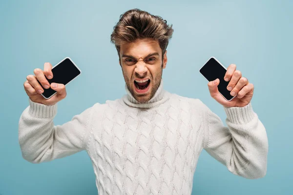 Homem agressivo gritando e segurando dois smartphones com telas em branco, isolado em azul — Fotografia de Stock