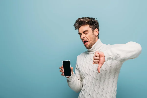 Unset emotional man showing thumb down and smartphone with blank screen, isolated on blue — Stock Photo