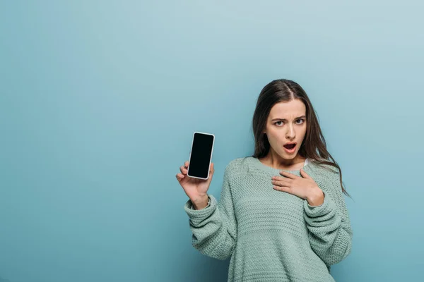 Mujer conmocionada mostrando teléfono inteligente con pantalla en blanco, aislado en azul - foto de stock