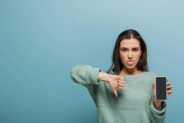 Mujer sacando la lengua mientras muestra el pulgar hacia abajo y el teléfono inteligente con pantalla en blanco, aislado en azul — Stock Photo