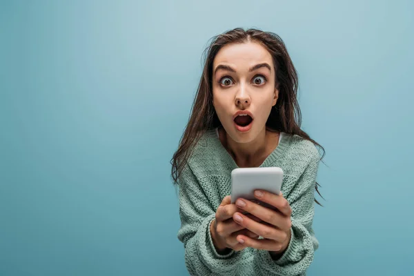 Schockierte Frau mit geöffnetem Mund mit Smartphone, isoliert auf blauem Grund — Stockfoto