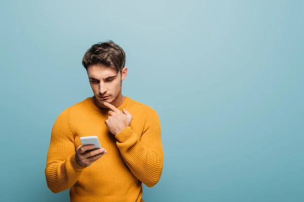 Hombre reflexivo guapo usando el teléfono inteligente, aislado en azul - foto de stock