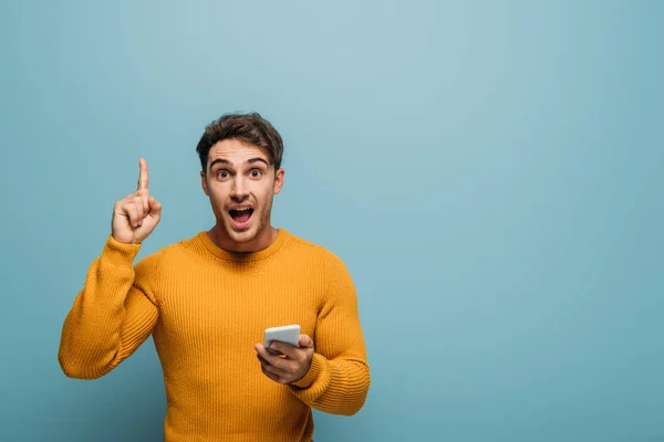 Homem surpreso usando smartphone e apontando para cima, isolado em azul — Fotografia de Stock