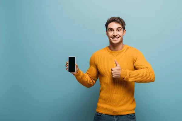 Felice uomo mostrando pollice in su e smartphone con schermo bianco, isolato su blu — Foto stock