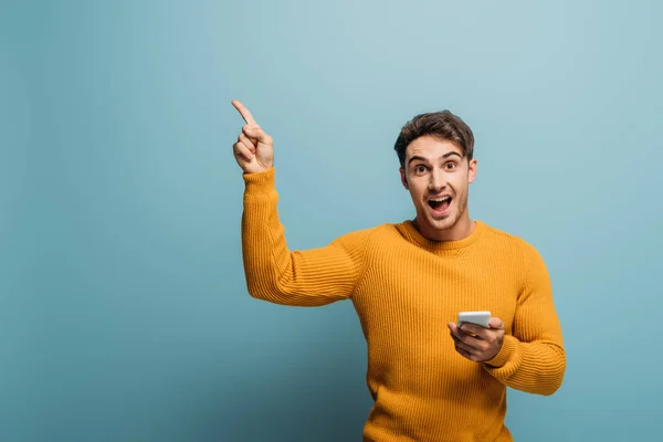 Hombre guapo excitado señalando y usando teléfono inteligente, aislado en azul - foto de stock
