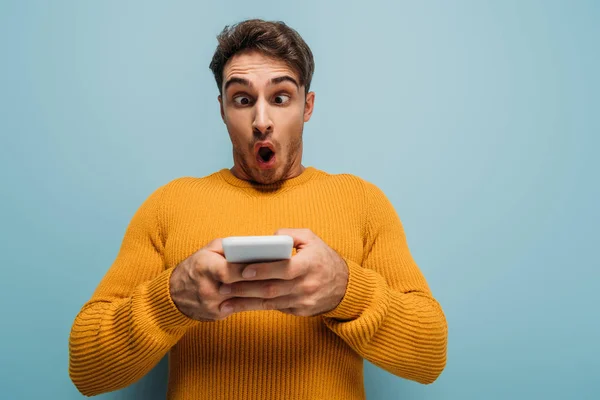 Sorprendido hombre usando teléfono inteligente, aislado en azul - foto de stock