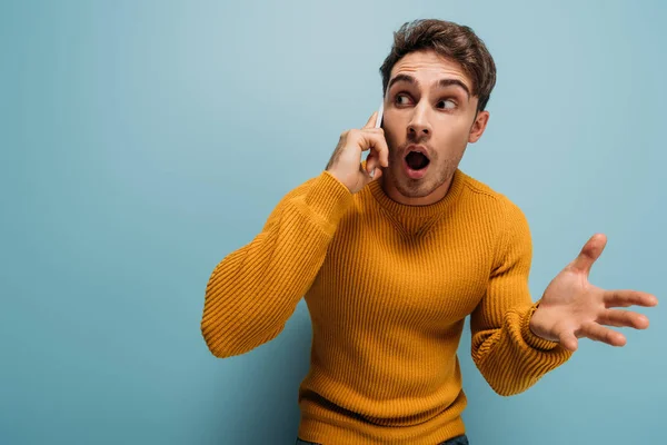 Sorprendido hombre guapo hablando en el teléfono inteligente, aislado en azul - foto de stock