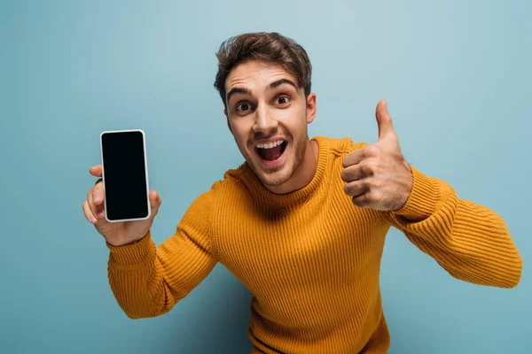 Aufgeregter junger Mann zeigt Daumen hoch und Smartphone mit leerem Bildschirm, isoliert auf blauem Grund — Stockfoto