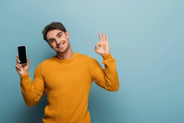 Hombre alegre mostrando signo ok y teléfono inteligente con pantalla en blanco, aislado en azul - foto de stock