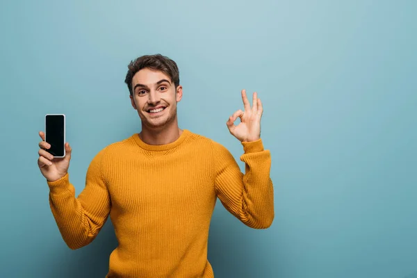 Positive man showing ok sign and smartphone with blank screen, isolated on blue — Stock Photo