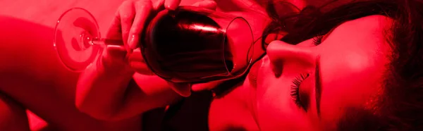 Plano panorámico de atractiva mujer seductora bebiendo vino en la luz roja - foto de stock