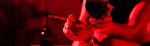 Visão parcial da mulher segurando cigarro e copo de vinho em luz vermelha, tiro panorâmico — Fotografia de Stock