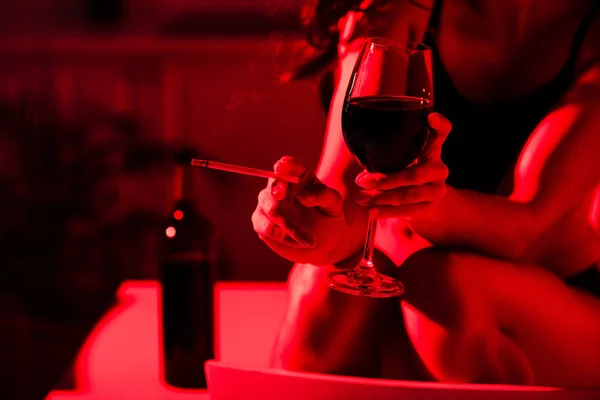 Vue recadrée de fille sexy tenant cigarette et verre de vin en lumière rouge — Photo de stock