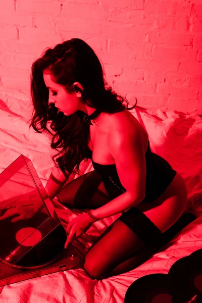 Atractiva chica seductora sentada en la cama con discos de vinilo en luz roja - foto de stock