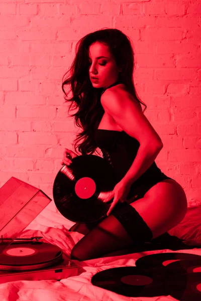 Attractive sexy young woman sitting on bed with vinyl records in red light — Stock Photo