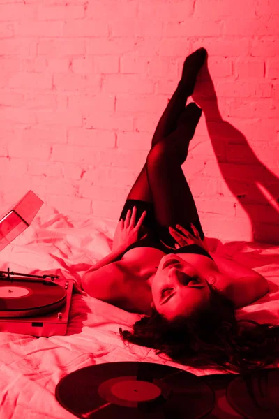 Attractive seductive girl lying on bed with vinyl records in red light — Stock Photo
