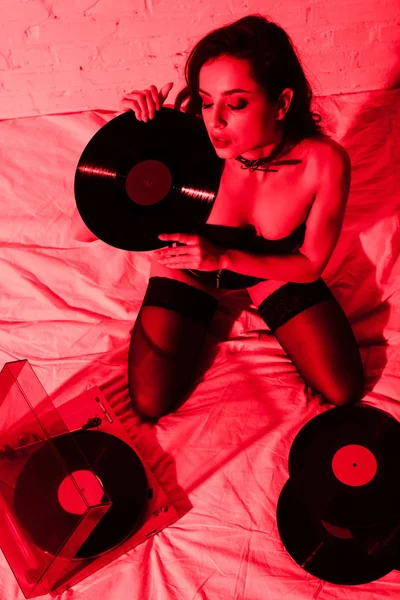 Mulher sexy em meias sentado na cama com discos de vinil em luz vermelha — Fotografia de Stock