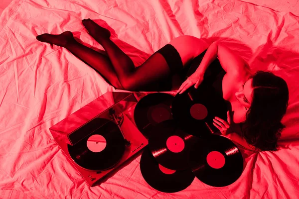 Belle femme sexy en bas couché sur le lit avec vinyle disques en lumière rouge — Photo de stock